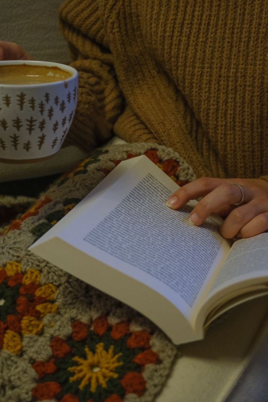 A cozy scene of a person holding a book and coffee mug, wrapped in a colorful blanket.